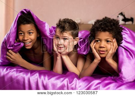 Three smiling boys under blanket. Children laying on silk bed. So nice to be together. Holiday morning at cousin's place.