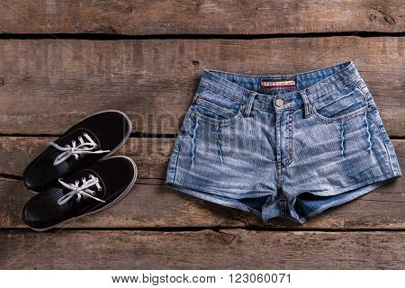 Denim shorts and black keds. Shorts and shoes on table. Simple clothing on old shelf. Classic clothes on aged floor.