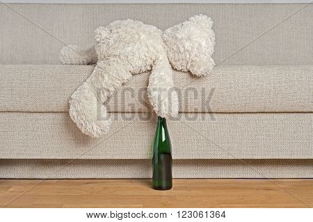 White Teddy bear is laying on edge of the sofa with the bottle in it's hand