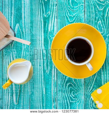 cup of black tea on yellow plate and yellow milk jug and sugar-bowl
on turquoise colored old wooden table with yellow napkin at polka dots top view