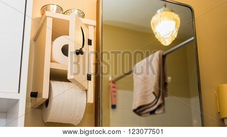 Wooden toilet paper holder with small door, crescent moon cut into door, toilet paper inside and one roll on spool.  Essential paper needs in bathroom.   Low angle partial view of bathroom, mirrored light, towel on a shower curtain bar.