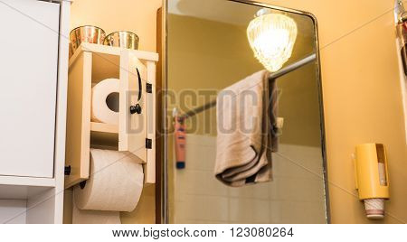 Wooden toilet paper holder with small door, crescent moon cut into door, toilet paper inside and one roll on spool.  Essential paper needs in bathroom.   Low angle partial view of bathroom, mirrored light, towel on a shower curtain bar.