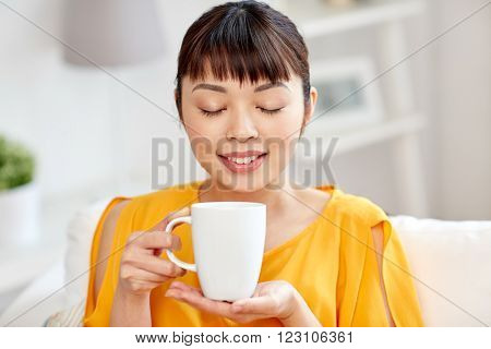 people, drinks and leisure concept - happy asian woman sitting on sofa and drinking tea from cup or mug at home