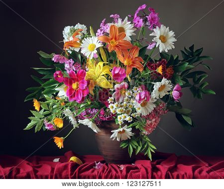 Still life with a bouquet of garden flowers in a clay jug on the table with a red tablecloth against a dark background.