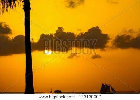 KEY WEST, USA - MAY 10, 2015: The sun going down above the Key West Bight on the luminous orange sky with some clouds and silhouettes of a sailing ship and a palm tree.