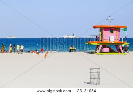 MIAMI BEACH, USA - MAY 9, 2015: A beach section with a pink lifeguard station and some people sitting or standing, a trash can in front and two cruise liners on the ocean in the back.