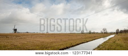 Picturesque Dutch polder landscape with a small stream diagonal across the image and yellowed grass on both sides of it. It is a cloudy day at the end of the winter season.