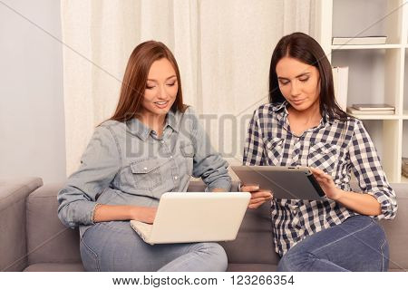 Portrait of two calm young woman holding laptop and tablert and reading news