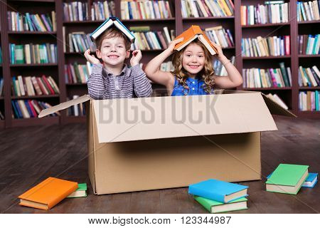 Playful childhood. Little children having fun with cardboard box. Children with books on heads. Children playing at room with bookshelf