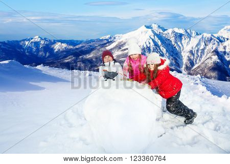 Three kids balling up the huge snowball making a snowman on top of beautiful mountain