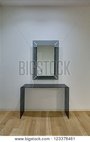 Light wall with mirror with frosted glass frame on it. Their is a frosted glass table under mirror. White door reflected in the mirror. On the floor there is a light parquet.