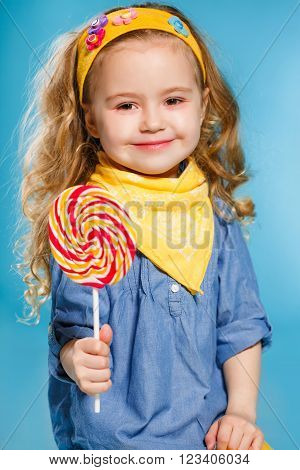 Funny little girl with long, curly hair,with a yellow handkerchief around her neck, a sweet smile,is dressed in a blue shirt and yellow pants,posing in Studio standing against a blue background, holding a large,round, colourful Lollipop
** Note: Soft Foc
