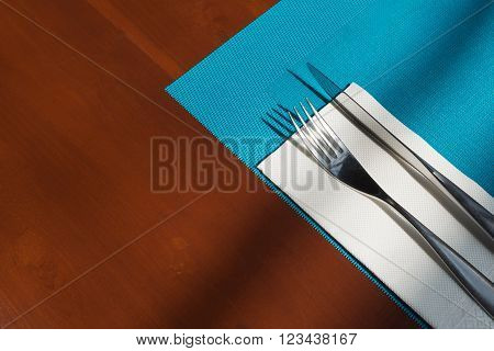 Silverware and tissue and blue mat napery on brown wooden table. Simple table setting in restaurant with light and shadow.