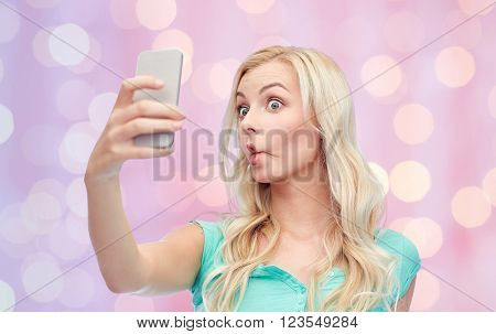 expressions, technology and people concept - funny young woman or teenage girl taking selfie with smartphone and making fish face over pink holidays lights background