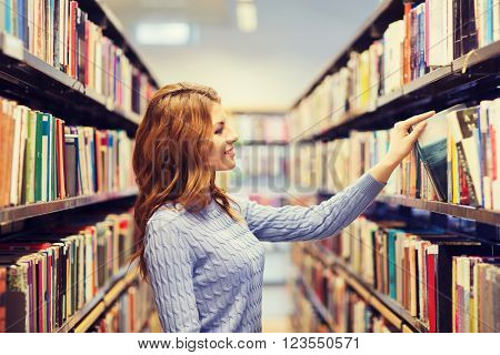 people, knowledge, education and school concept - happy student girl or young woman taking book from shelf in library