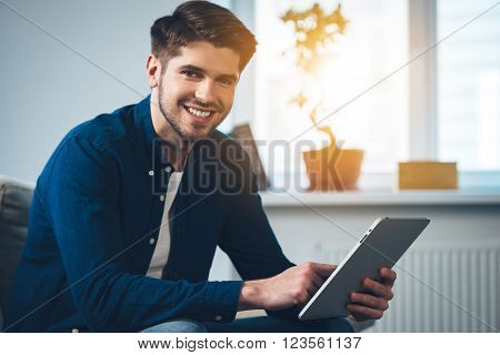 This tablet exceeds his expectations. Side view of handsome young man using his digital tablet and looking at camera with smile while sitting on the couch at home