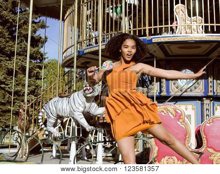 cool real modern teenage girl with candy near carousels at amusement park walking, having fun
