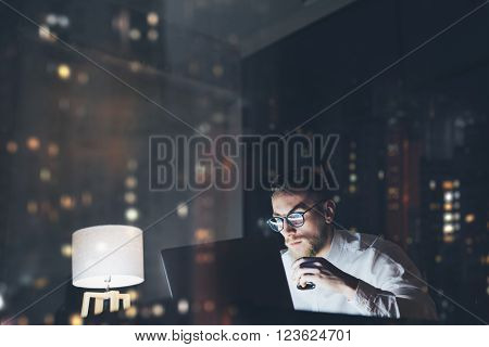 Bearded young businessman working on modern loft office at night. Man using contemporary notebook texting message, holding cup espresso, blurred background, bokeh