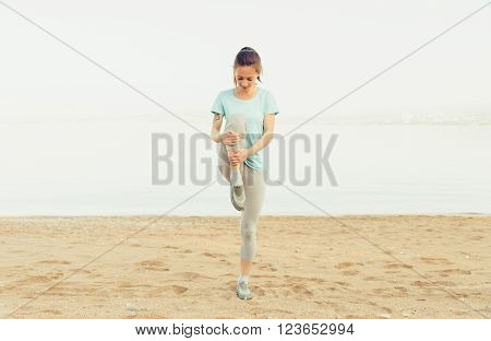 Sporty young woman stretching her legs and preparing to run on sand beach in summer workout. Concept of healthy lifestyle