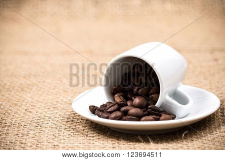 White coffeecup and plate with spilled coffeebeans on gunny textile