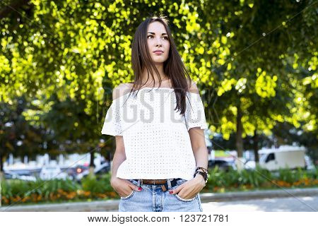A beautiful girl standing in a park, in a white jacket, fashion style urban life, a bright sunny summer day. Outdoors brunette woman.