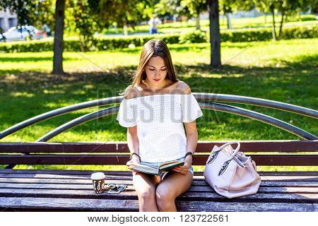 Young girl student sitting on a park bench, reading a paper book with a cup of coffee or a tea bag in a white jacket, a bright summer day outdoors, fashion lifestyle.