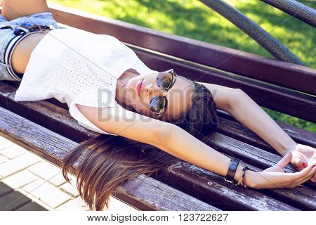 Young modern student beautiful girl, brunette lying on a park bench relaxing in the fresh air. Close-up. Fashion style glamorous woman, concept idea long hair look on the bench stretches.
