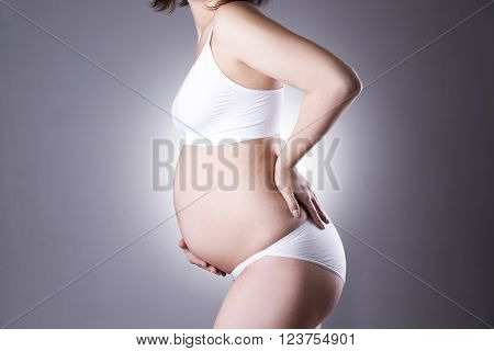 Caucasian pregnant woman in white lingerie with back pain on gray studio background. Pregnancy concept