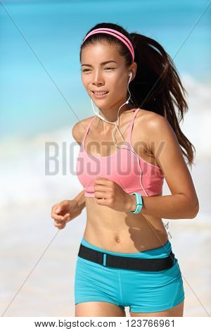 Happy running woman listening to music with earphones and fitness watch activity tracker in sportswear and sports bra. Healthy Asian runner girl on beach with wearable tech gear.
