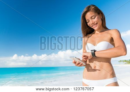 Bikini woman using smart phone syncing watch data for smartwatch app on smartphone. Happy Asian girl on beach summer holidays in tropical destination looking at screen of activity tracker device.