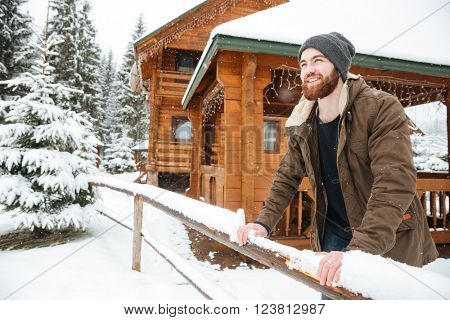 Portrait of cheerful handsome bearded man standing in front of wooden cottage in winter forest