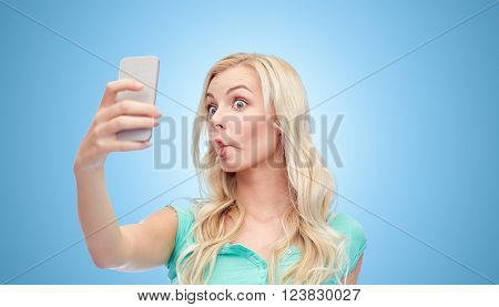 expressions, technology and people concept - funny young woman or teenage girl taking selfie with smartphone and making fish face over blue background