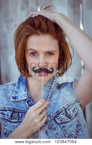 beautiful hipster woman holding a fake mustache to her face, against a wooden backhround