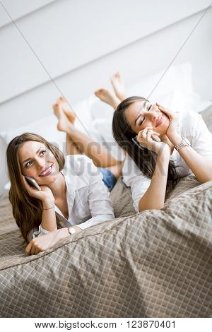 Two beautiful girls talking on the phone and smiling while lying on a luxorious bed
