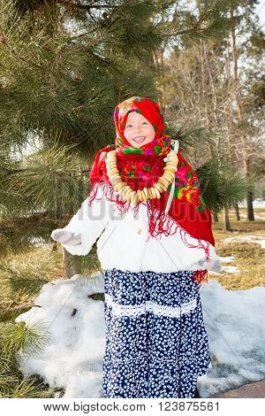 Child girl in Russian pavloposadskie folk scarf on head with floral print and with bunch of bagels on background of snow. Portrait of girl dressed in Russian style