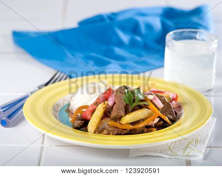 Beef and onion stir fry, lomo saltado, a typical Peruvian dish served in yellow dish on white tile counter top with blue napkin