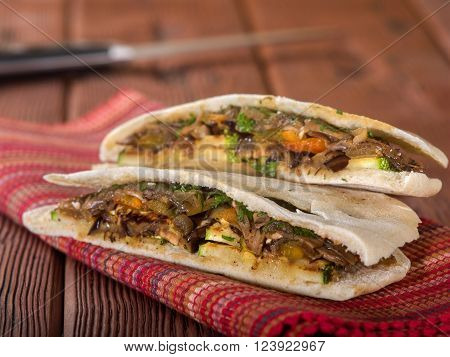 Vegetarian pita bread sandwich with eggplant and zucchini sitting on red textural place mat on wooden table top