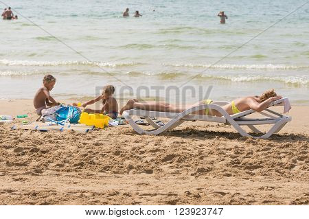 On The Beach Near The Water With A Beach Chair Sunbathing Woman Near The Children Playing In The San