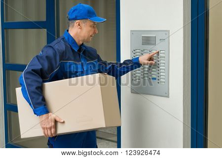 Delivery Man With Cardboard Box Pressing Button Of Intercom To Enter Building