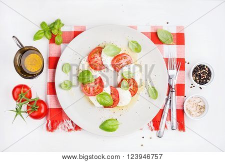 CLassic Italian Caprese salad with tomatoes, mozzarella di Buffala and fresh basil on red and white checkered napkin over white wooden background. Top view