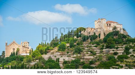 Pilgrimage church of Sant Salvador on green hill in Arta village, Mallorca island, Spain. Beautiful landsacape with medieval architecture, green trees and blue sky with clouds on a sunny day.