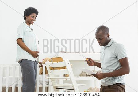 Pregnant woman looking at man reading a list while fixing a baby chair at home