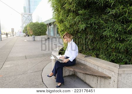 Female statistician working with color diagrams and laptop outdoors near green plant. Concept of working with theoretical or applied statistics. Young woman wears business clothes.