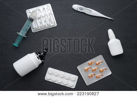 Thermometer, nasal drops tablets and ampoules with medicine on dark slate board. Flat lay, copy space. Meds for flue treatment and antibiotics, health care concept