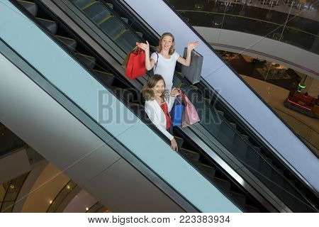 Portrait of two playful beautiful women looking at camera, standing on escalator and holding paper-bags in mall. Shopping concept. Side view.