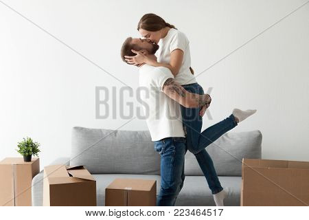 Happy loving couple kissing feeling excited about moving into new home, man lifting holding woman in hands standing among cardboard boxes, glad tenants enjoying relocation, starting living together