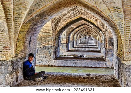 Isfahan, Iran - April 24, 2017: The arched line under the bridge Allahverdi Khan above the river Zayandeh, one young man sits on a stone slab, cross-legged.