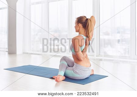 Beautiful young woman practices yoga asana Gomukhasana - Cow face pose at the bright yoga class with large windows