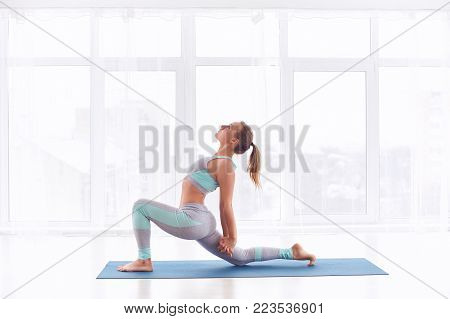 Beautiful sporty fit yogini woman practices yoga asana Ashva Sanchalasana - equestrian pose at the bright yoga class with large windows