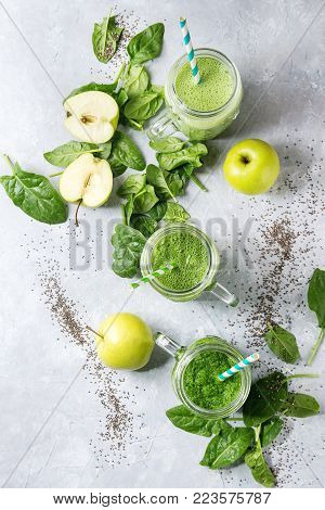 Variety of three color green spinach kale apple yogurt smoothie in mason jars with retro cocktail tubes and ingredients above over gray texture background. Healthy vegan detox eating. Top view, space